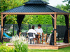 Two people sitting at a table under a wooden gazebo with black roof and curtains in a garden setting.